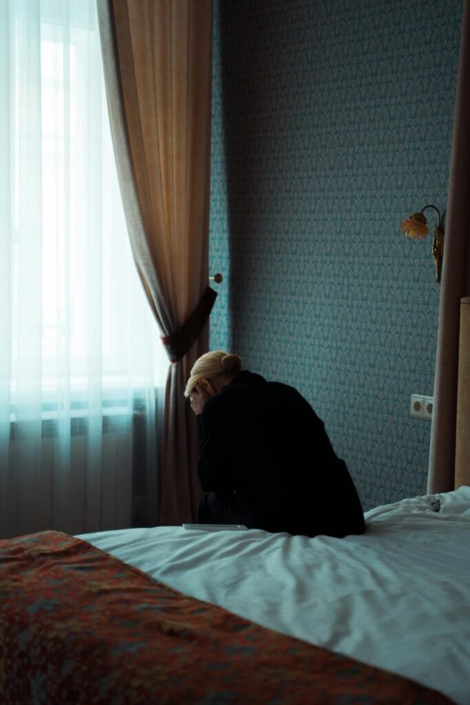 A solitary woman sits on a bed, gazing out a window, evoking feelings of solitude and introspection.